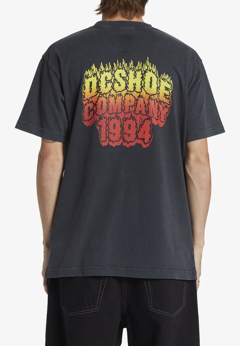 T-shirt oversize gris foncé avec un motif de flammes vives rouges, jaunes et oranges, et "DC Shoe Company 1994" imprimé au dos.