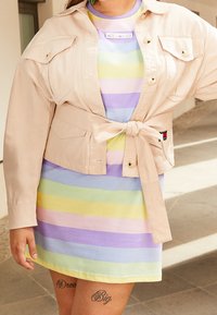 Veste beige courte avec poches, nouée à la taille, portée sur une robe rayée multicolore dans des teintes pastel de violet, jaune et vert.