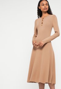 Hosszú ujjú, bordázott ruha világos bézs színben, körszerű szoknyával. Három díszítő gomb található a nyak részén. Simá szövet és testhez simuló sziluett jellemzi.