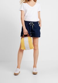 Sac seau tissé jaune et blanc avec une poignée en bois, associé à un t-shirt blanc à col en V, un short marine et des baskets blanches avec des semelles en jute.