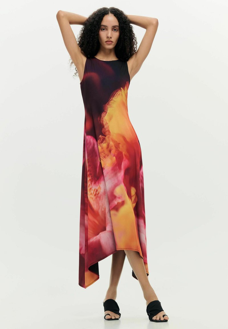 Kvinde med krøllet hår iført en ærmeløs, asymmetrisk kjole med røde, orange og pink blomsterprint, stående med armene hævet.