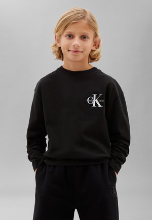 Niño rubio con ojos azules que lleva una sudadera negra de Calvin Klein y pantalones negros a juego, de pie con las manos en los bolsillos sobre un fondo gris.