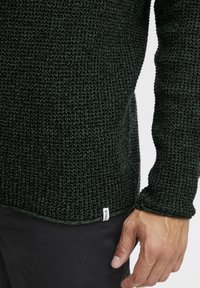 Dark green knit sweater with a textured pattern, featuring a rounded hem and a small label at the bottom. Fitted sleeve cuffs.