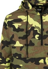 Kamouflaged hoodie i gröna och gula nyanser, med halvdragkedja, dragsko på huvan och en framficka. Tillverkad av lättviktsmaterial.