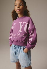 Felpa viola con scritte bianche, polsini a costine e orlo elastico, abbinata a pantaloni corti di jeans chiari con vita elastica e tasche.