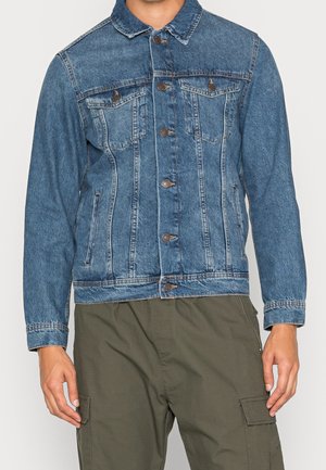 Geknöpfte blaue Jeansjacke mit Brust- und Seitentaschen, getragen über olivgrünen Cargohosen.