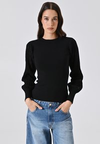 Schwarzer Pullover aus Strickmaterial mit strukturierten, puffigen Ärmeln und taillierter Passform. Kombiniert mit hellblauen, hochgeschnittenen Jeans.