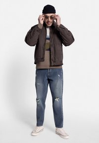 Barna vadászdzseki, bézs mintás pulóver, kopott kék farmernadrág és fehér tornacipő. Kiegészítők: napszemüveg és tengerészkék sapka.