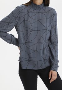 Lyseblå bluse med høj krave og lange ærmer. Har et geometrisk mønster i mørkeblå, tekstureret stof, og løs pasform.
