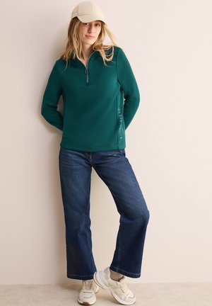 Teal fleece pullover met rits en de tekst "FOCUS AHEAD" aan de zijkant, gecombineerd met blauwe rechte jeans en witte sneakers.
