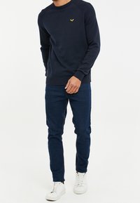 Marineblauwe trui met een geribbelde schouderontwerp, voorzien van een klein geel logo op de borst. Gepaard met donkerblauwe jeans en witte sneakers.