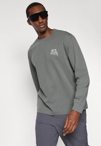 Camisola técnica cinza de mangas longas com um gráfico branco no lado esquerdo do peito. Combinada com calças cinzas escuras e óculos de sol pretos oversized.