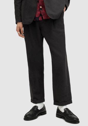 Veste de costume noire par-dessus une chemise à fleurs rouges, pantalon large noir et mocassins noirs avec des chaussettes blanches. Tissu lisse et coupe moderne.