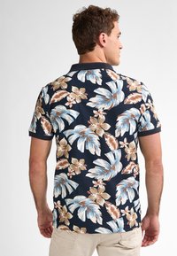 Polo de manga corta con fondo azul marino y grandes patrones de flores y hojas en tonos beige, azul y marrón, tejido texturizado.