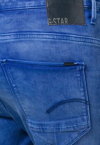 Blå jeans i denim med en bakficka som har en kurvad sömdetalj och en läderetikett präglad med "G.STAR." Texturerad tyg och slät yta.