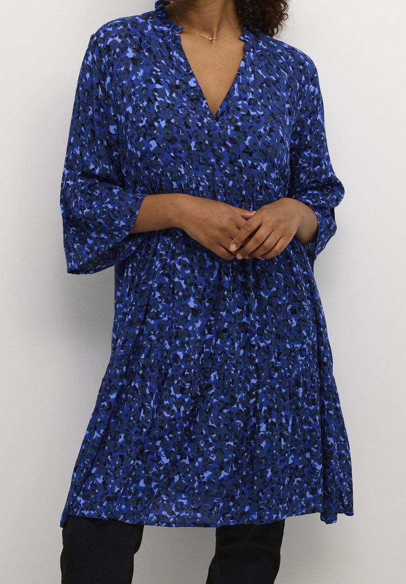 Femme portant une robe bleue à fleurs longueur genoux, manches trois-quarts et col en V, debout les mains légèrement jointes.