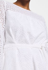 Vestido blanco con una tela texturizada que presenta patrones de ojales. Las mangas largas y un lazo en la cintura acentúan el corte. Escote redondeado.