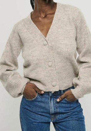 Person trägt eine beige, gerippte Strickjacke mit großen Knöpfen und blaue, hoch taillierte Jeans, die Hände in den vorderen Taschen vergraben.