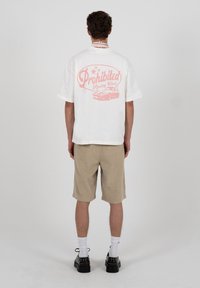 Άνδρας που φοράει λευκό υπερμεγέθες T-shirt με ροζ γράμματα "Prohibited Racing Club" και γραφικό αυτοκινήτου στην πλάτη, μπεζ σορτς, λευκές κάλτσες, μαύρα παπούτσια, όρθιος.