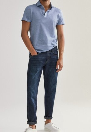 Man wearing light blue short-sleeve polo shirt, dark blue rolled-up jeans, and white sneakers, standing with one hand in pocket.