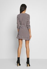 Nainen seisoo, päällään pitkähihaista houndstooth-kuosisella mini-mekolla ja mustilla korkokengillä nilkan korkeudelta, kuvattu takaa.