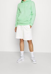 Grön huvtröja med ficka fram, ihop med vita shorts. Vita sneakers med grå detaljer och matchande vita strumpor. Enfärgad bakgrund.