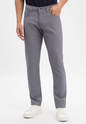 Mann trägt graue, schmal geschnittene Hose und weiße Turnschuhe, eine Hand in der Tasche, steht vor einem schlichten hellen Hintergrund.