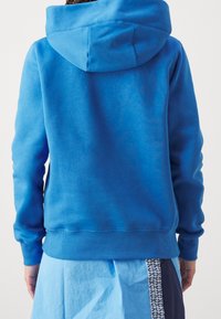 Blå huoodtröja med känguruficka, ribbade ärmslut och nederkant. Mjuk textur och avslappnad passform. Synliga sömmar vid halsringningen.