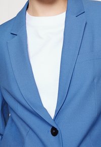 Jasnoniebieska marynarka wykonana z fakturowanej tkaniny, z wykładanymi klapami, jednym czarnym guzikiem oraz białą koszulką pod spodem.