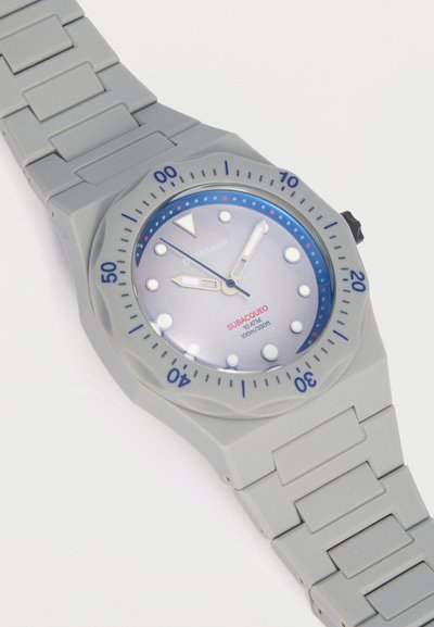 Γκρι καταδυτικό ρολόι με μπλε και λευκή στεφάνη, φωτεινά δείκτες, κυκλικούς δείκτες ώρας και λουράκι από καουτσούκ, με την ένδειξη «SUBACQUEO 10 ATM 100m/330ft».