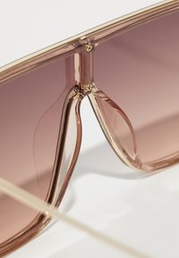 Nærbilde av transparente rosa solbriller med gullskruer og gradient lilla linser på hvit bakgrunn.