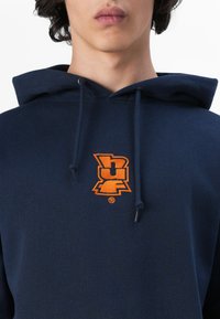 Sweatshirt azul-marinho feito de algodão, com um bolso frontal tipo canguru, apresentando um logótipo bordado laranja no peito e cordões para o capuz.