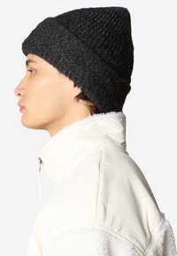Berretto lavorato a maglia nero con un motivo texture, indossato con una giacca crema con zip e colletto soffice in tessuto liscio.