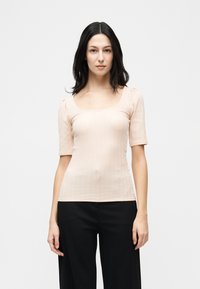 Γυναίκα με μακριά σκουρόχρωμα μαλλιά, ντυμένη με ροζ ανοιχτό ριμπ μπλούζα με κοντά μανίκια και μαύρο παντελόνι, στέκεται μπροστά σε απλό λευκό φόντο.