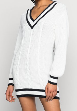 Kobieta w białej sukience swetrowej z warkoczowym splotem, z czarnymi paskami na dekolcie w kształcie litery V, mankietach i dolnym brzegu, stojąca na jasnym tle.