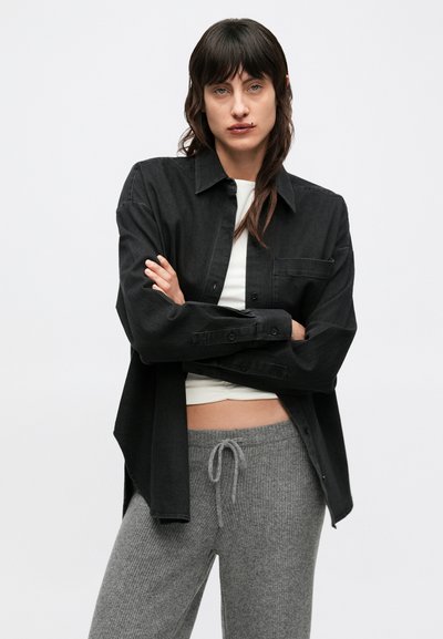 Czarna koszula na guziki z kieszenią na piersi, noszona na białej bluzce, zestawiona z szarymi prążkowanymi spodniach dresowych z wiązanym pasem.