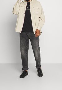 Camisa botón de color beige claro sobre una camiseta negra, combinada con jeans gris oscuro y zapatos negros de cordones. Presenta bolsillos en el pecho y un logotipo bordado.