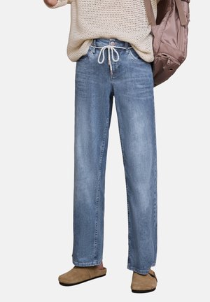 Osoba nosząca jasnoniebieskie szerokie dżinsy ze ściągaczem w talii, beżowy sweter z dzianiny, brązowe wsuwane buty i niosąca lawendowy plecak.