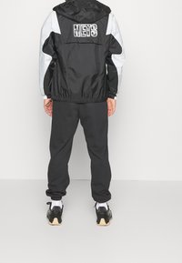 Sort vindjakke med grå accenter, hætte og "NETS" logo på ryggen. Sammen med sorte sweatpants og sorte sneakers med en lys sål.