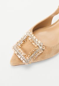 Chausson beige en daim à talon slingback avec un bout pointu, orné d'une boucle carrée en strass dans une palette de couleurs or et transparente.