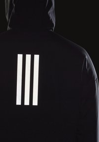 Personne portant une veste à capuche foncée avec trois bandes blanches réfléchissantes verticales dans le dos sur un fond noir.