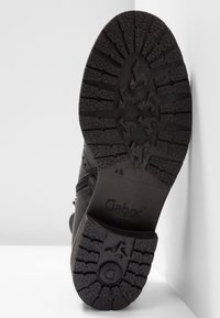 Semelle d'une chaussure noire Gabor avec des motifs de profil profond et une adhérence texturée, inclinée verticalement contre une surface blanche d'angle.