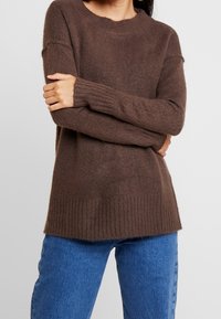 Hnědý pletený svetr s žebrovaným lemem a manžetami, kulatým výstřihem a mírně oversize střihem, spárovaný s modrými džíny.