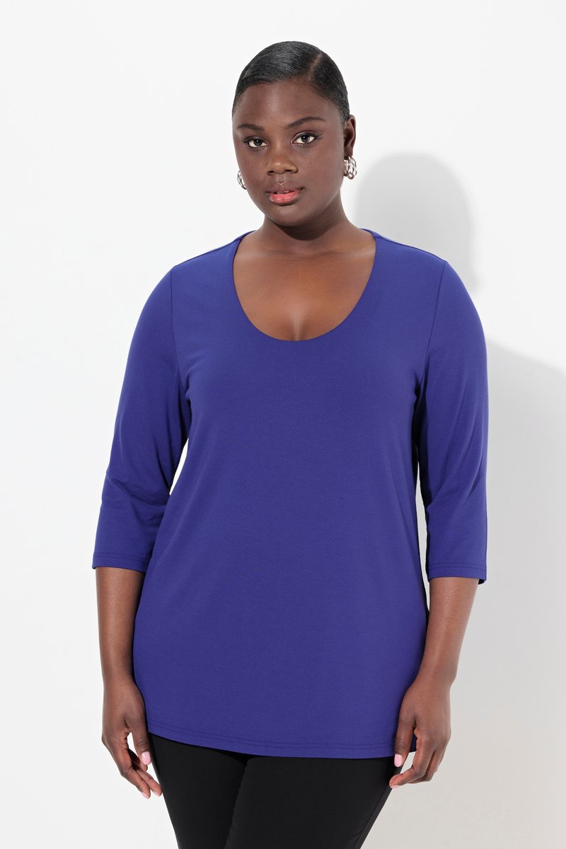 Femme à la peau foncée et aux cheveux courts portant un haut violet ajusté à col rond avec des manches trois-quarts et un pantalon noir, debout devant un fond blanc.