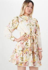 Giovane donna che indossa un vestito floreale a maniche lunghe con una cintura in vita e dettagli ad occhiello, in piedi con una mano sui fianchi.