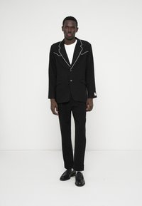 Fato preto com bolinhas brancas, apresentando um blazer de um só botão com piping branco e calças a condizer. Inclui uma t-shirt branca simples por baixo.