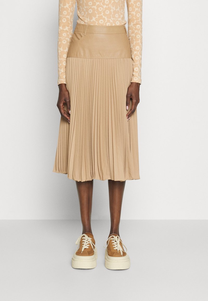 Beige plisserad kjol med en åtsittande midja i syntetiskt läder, parat med bruna sneakers med strukturerat ovandel och tjock vit sula.