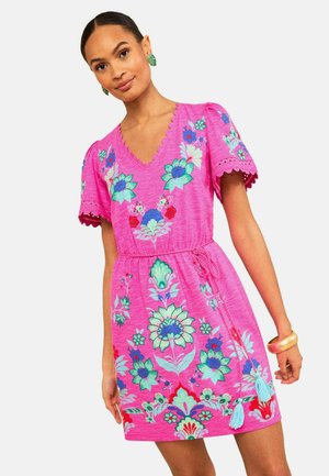 Kobieta w jasnoróżowej kwiecistej sukience z falistymi brzegami, krótkimi rękawami, dekoltem w serek i przewiązanym paskiem w talii z frędzlami.