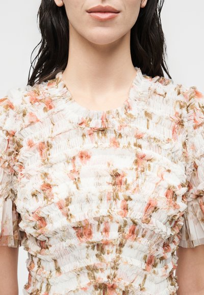 Γυναίκα φοράει ένα λευκό υφασμάτινο τοπ με στρωτές λουλουδάτες βολάν σε ροζ και πράσινους τόνους, κοντά μανίκια και στρογγυλή λαιμόκοψη.