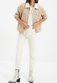 Beige sako s bílým umělým kožešinovým límcem, v kombinaci s bílými kalhotami s vysokým pasem a kotníkovými botami s puntíky, metalickým špičkou a širokým podpatkem.